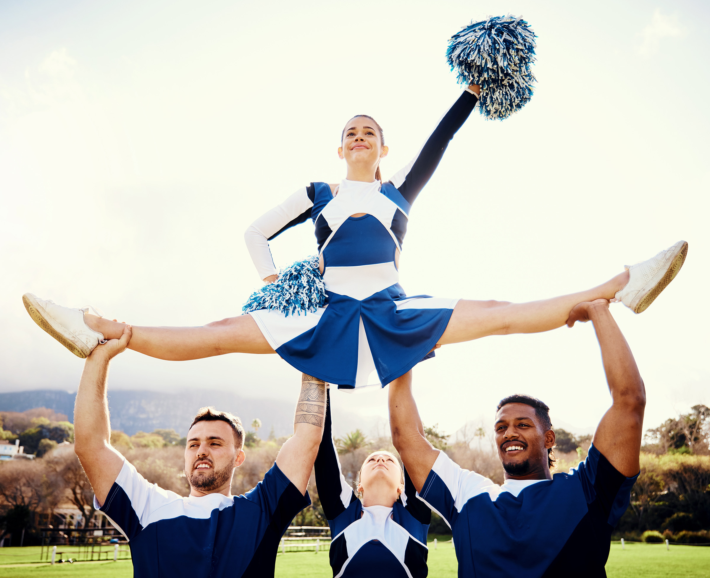 A group of cheerful people in blue and white outfits are performing a cheerleading stunt in a sunny outdoor setting. One cheerleader is held aloft, doing the splits.